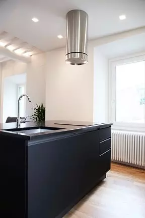 Questa cucina moderna presenta un design elegante e funzionale. L'isola centrale in laminato nero ospita il piano cottura con cappa cilindrica in acciaio, creando un punto focale nella stanza. Le colonne forno e frigo, anch'esse in laminato nero, si affiancano all'isola creando un'armonia di volumi e colori. Il pavimento in parquet di rovere chiaro aggiunge un tocco di calore e luminosità all'ambiente. Questa cucina è un esempio perfetto del lavoro di un designer di interni esperto che ha saputo combinare stile e funzionalità per creare un ambiente elegante e confortevole.