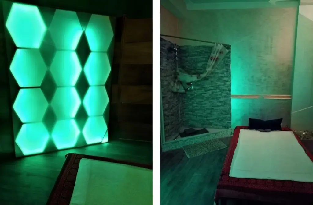 Immagine divisa in due che mostra l'interno di una moderna spa illuminata da luci a LED verdi. La sezione sinistra presenta una grande parete decorativa composta da esagoni retroilluminati con LED RGB che cambiano colore (attualmente verdi), posizionata dietro una panca con rivestimento. La sezione destra mostra una zona con parete in pietra, una doccia con tenda e un lettino da massaggio preparato con lenzuola bianche e un cuscino scuro, il tutto immerso nella stessa luce ambientale verde.