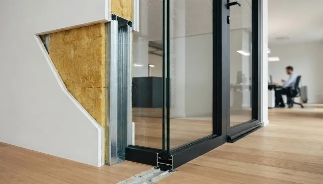 Immagine ravvicinata e fotorealistica che mostra una sezione costruttiva per l'isolamento acustico in un ufficio. A sinistra, una controparete a secco aperta rivela l'intercapedine con lana di roccia gialla tra profili metallici e lastre di cartongesso bianco. Al centro, l'attacco a pavimento e soffitto di una partizione in vetro stratificato con profilo in alluminio nero e giunto tecnico nel parquet in rovere. Sulla destra sullo sfondo, si intravede un ambiente d'ufficio con una scrivania.